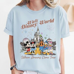 May include: Light blue t-shirt featuring a graphic of Disney characters in front of a castle. The text "Walt Disney World" and "Where Dreams Come True" are printed above and below the graphic. The shirt has short sleeves.