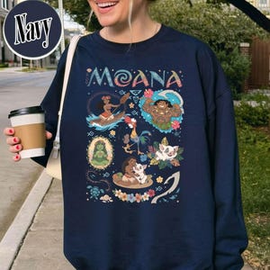 May include: Navy blue sweatshirt with the word "Moana" in a decorative font. The design features cartoon illustrations of characters and scenes from the film, including a woman in a canoe, a muscular man, and animals.