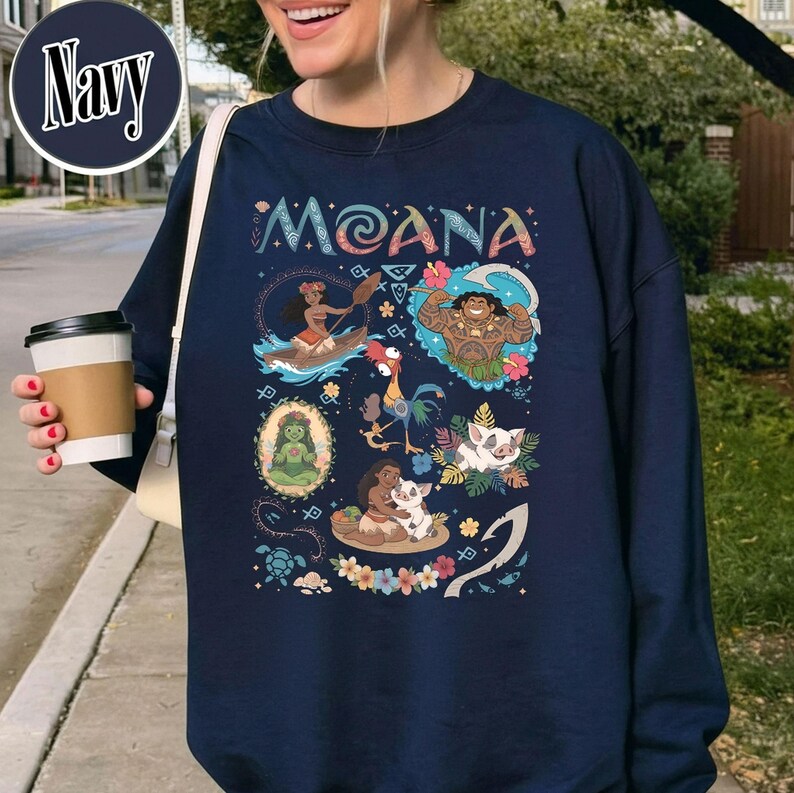 May include: Navy blue sweatshirt with the word "Moana" in a decorative font. The design features cartoon illustrations of characters and scenes from the film, including a woman in a canoe, a muscular man, and animals.