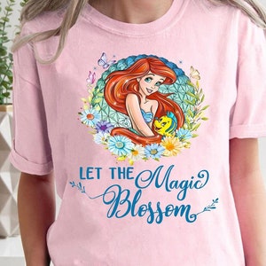 May include: A light pink t-shirt featuring a colorful graphic of a mermaid with red hair, a blue seashell bra, and a yellow fish. The text "LET THE Magic BLOSSOM" is written in blue script below the graphic.