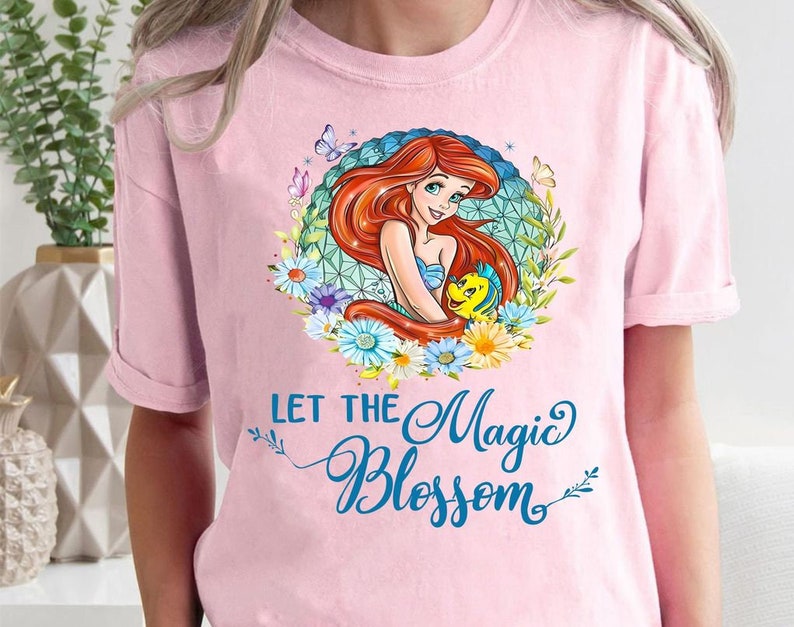 May include: A light pink t-shirt featuring a colorful graphic of a mermaid with red hair, a blue seashell bra, and a yellow fish. The text "LET THE Magic BLOSSOM" is written in blue script below the graphic.