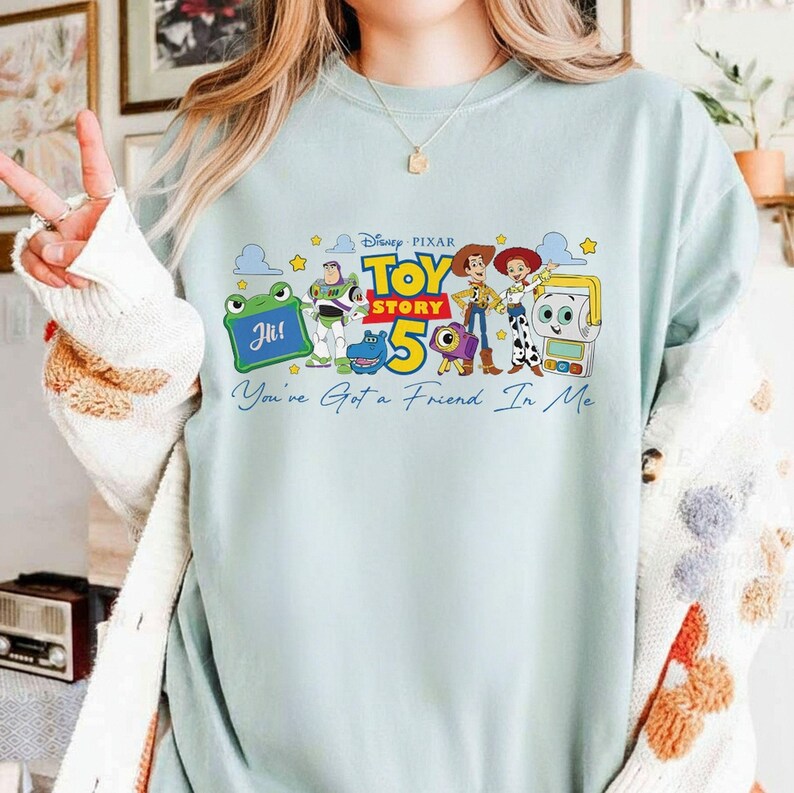 May include: Light blue sweatshirt featuring a colorful graphic of characters from the Disney Pixar movie Toy Story 5. The design includes Buzz Lightyear, Woody, and other characters, with the text "Toy Story 5" and "You've Got a Friend In Me".