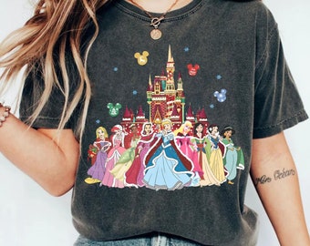 Könnte beinhalten: Ein dunkelgraues T-Shirt mit einem weihnachtlichen Design, das Disney-Prinzessinnen und das Schloss von Aschenputtel zeigt. Die Prinzessinnen tragen winterliche Outfits und stehen vor dem Schloss. Das Schloss ist mit Weihnachtslichtern und Ornamenten geschmückt.