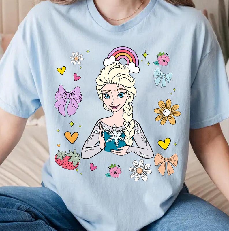 May include: A light blue t-shirt with a cartoon image of Elsa from Frozen. The design includes a rainbow, flowers, hearts, and a snowflake.