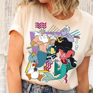 May include: A cream-colored t-shirt with a graphic print of characters from the Disney movie The Little Mermaid. The graphic features King Triton, Ariel, Ursula, and Flounder in a colorful, geometric design.