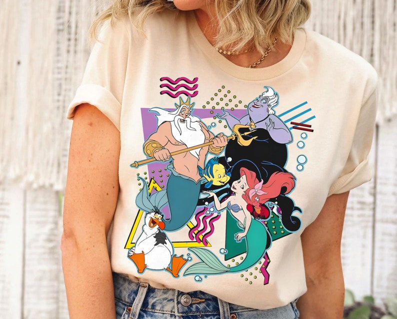 May include: A cream-colored t-shirt with a graphic print of characters from the Disney movie The Little Mermaid. The graphic features King Triton, Ariel, Ursula, and Flounder in a colorful, geometric design.
