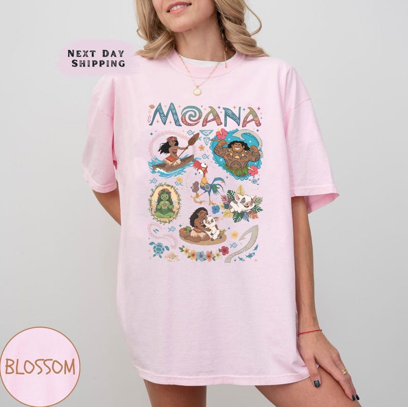 May include: A light pink t-shirt with the word "MOANA" in colourful letters. The shirt features cartoon illustrations of characters from the film. The t-shirt has a comfortable, oversized fit.