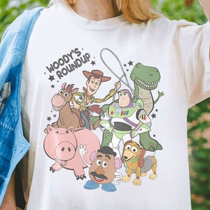 Puede incluir: Camiseta de color crema con un gr&aacute;fico colorido de los personajes de Toy Story. El dise&ntilde;o incluye a Woody, Buzz Lightyear, Rex, Hamm, Mr. Potato Head, Slinky Dog y un caballo. El texto "Woody's Roundup" est&aacute; encima de los personajes.