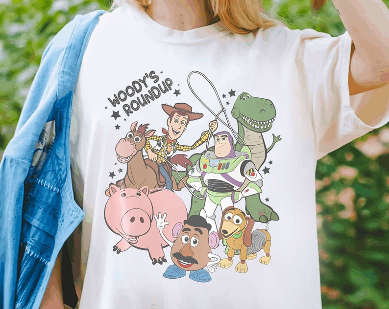 Puede incluir: Camiseta de color crema con un gr&aacute;fico colorido de los personajes de Toy Story. El dise&ntilde;o incluye a Woody, Buzz Lightyear, Rex, Hamm, Mr. Potato Head, Slinky Dog y un caballo. El texto "Woody's Roundup" est&aacute; encima de los personajes.