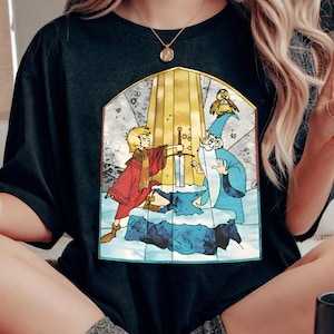 Op de afbeelding: Zwart T-shirt met een grafische print in de stijl van een glas-in-loodraam van een jonge jongen die een zwaard uit een steen trekt. De afbeelding bevat ook een tovenaar en een kleine vogel.