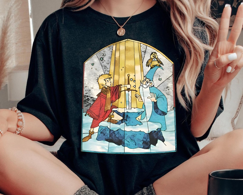 Op de afbeelding: Zwart T-shirt met een grafische print in de stijl van een glas-in-loodraam van een jonge jongen die een zwaard uit een steen trekt. De afbeelding bevat ook een tovenaar en een kleine vogel.