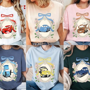 Op de afbeelding: Zes verschillende T-shirts met een cartoonauto in een parelmoerachtig frame met bloemmotieven. De auto's zijn rood, blauw, geel en bruin. De shirts zijn wit, roze, lichtblauw, donkerblauw en groen.