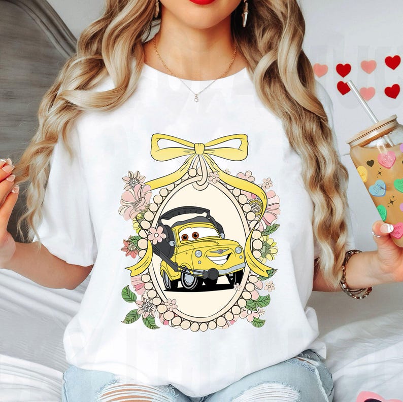 Op de afbeelding: Wit T-shirt met een gele cartoonauto in een bloemig frame. De auto heeft een glimlachend gezicht en is omringd door roze en witte bloemen.