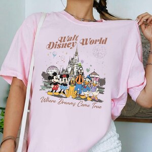 May include: A light pink t-shirt featuring a graphic of Disney characters, including Mickey Mouse, Minnie Mouse, Goofy, Donald Duck, and Daisy Duck, in front of a castle. The text "Walt Disney World" and "Where Dreams Come True" are printed above and below the characters.