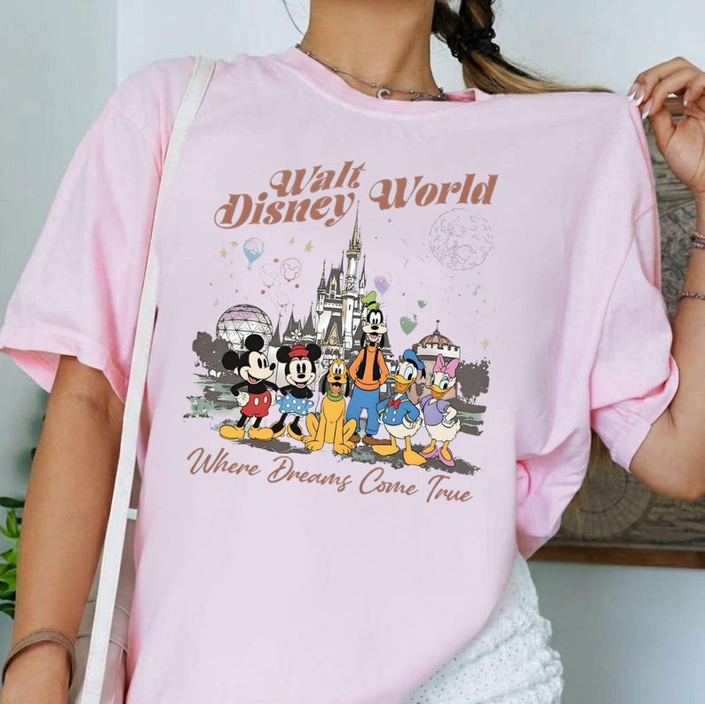 May include: A light pink t-shirt featuring a graphic of Disney characters, including Mickey Mouse, Minnie Mouse, Goofy, Donald Duck, and Daisy Duck, in front of a castle. The text "Walt Disney World" and "Where Dreams Come True" are printed above and below the characters.