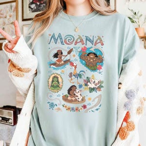 May include: Pale blue sweatshirt with the word "MOANA" in colourful lettering. The design includes cartoon illustrations of characters from the film, such as a woman in a canoe, a muscular man, and a pig.