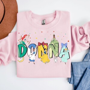 May include: Pink sweatshirt featuring a graphic of the name "DONNA" in green letters with Disney princesses in each letter. The princesses are Ariel, Belle, Snow White, Cinderella, and Tiana.