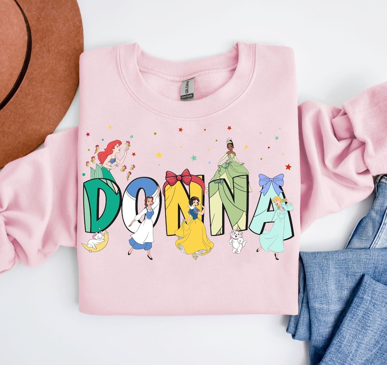 May include: Pink sweatshirt featuring a graphic of the name "DONNA" in green letters with Disney princesses in each letter. The princesses are Ariel, Belle, Snow White, Cinderella, and Tiana.