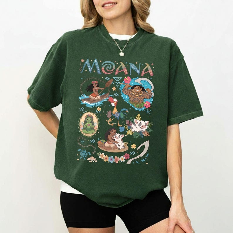 May include: Dark green t-shirt with the word "MOANA" in colourful letters. The shirt features cartoon characters and scenes from the film, including Moana in a canoe, Maui, Hei Hei, and Pua.