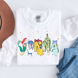 May include: White sweatshirt with a colourful design featuring Disney princesses spelling out the name "JONNA".