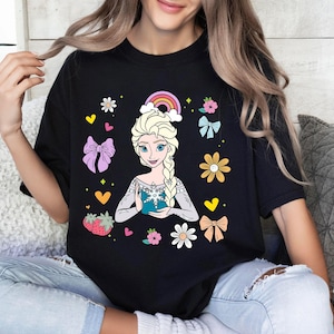 May include: Black t-shirt with a colorful graphic of a cartoon princess holding a snowflake. The graphic includes a rainbow, flowers, hearts, and bows.