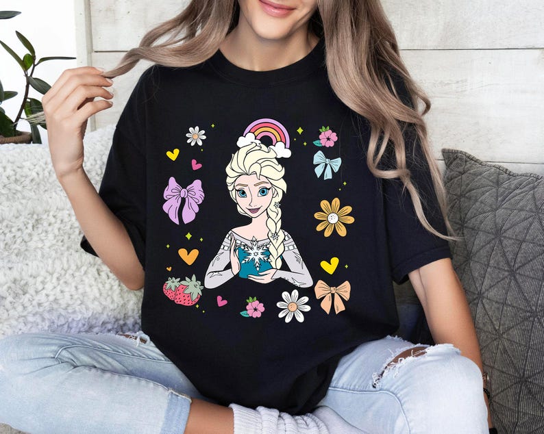 May include: Black t-shirt with a colorful graphic of a cartoon princess holding a snowflake. The graphic includes a rainbow, flowers, hearts, and bows.