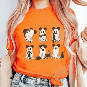 May include: Orange t-shirt featuring a design of six ghosts wearing Mickey Mouse ears and holding black cats. The ghosts are white with black eyes and mouths.