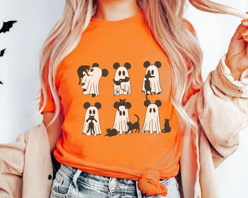 May include: Orange t-shirt featuring a design of six ghosts wearing Mickey Mouse ears and holding black cats. The ghosts are white with black eyes and mouths.