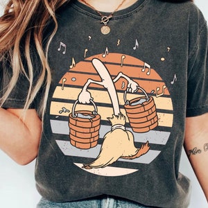 May include: A dark grey t-shirt featuring a vintage-style graphic of a broom holding two buckets. The graphic includes a sunset with orange, yellow, and red stripes and musical notes.