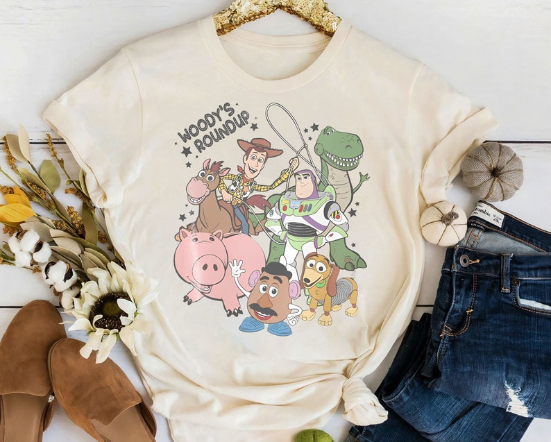 Puede incluir: Camiseta de color crema con un gr&aacute;fico de personajes de la pel&iacute;cula animada Toy Story. El dise&ntilde;o incluye a Woody, Buzz Lightyear, Rex, Hamm, Sr. Potato, Slinky Dog y Bullseye, con el texto "Woody's Roundup."