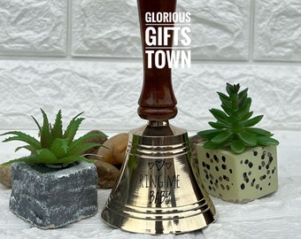 Könnte beinhalten: Eine goldfarbene Handglocke mit einem Holzgriff. Die Glocke trägt die Gravur "Ring Me Baby" mit drei Herzen über dem Text.