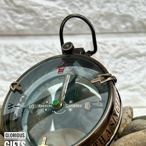 Może przedstawiać: Zabytkowy mosiężny kompas ze szklaną tarczą, z oznaczeniami kierunków i igłą. Kompas ma pętlę do zawieszania i jest otoczony małymi kamieniami. Widoczny jest tekst "GLORIOUS GIFTS TOWN".