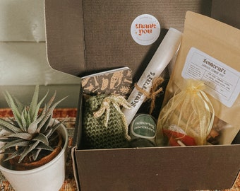 May include: A brown cardboard box filled with a variety of items, including a small green crocheted bag, a rolled up piece of paper with the word "SOUL" written on it, a small glass bottle with a label that says "Morning Incense", a small bag of dried herbs, and a small piece of paper with a thank you message. The box is sitting on a colorful rug.