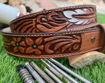 May include: Two brown leather belts with floral and feather tooling. The top belt features a feather design with white accents, while the bottom belt has turquoise flower accents. The belts are on green grass with leatherworking tools.