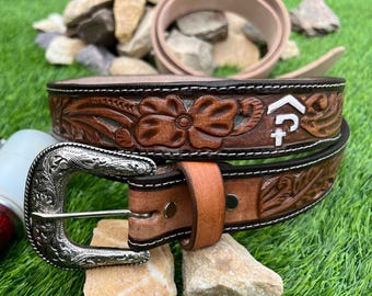 May include: A brown leather belt with a silver buckle. The belt has floral and geometric carved patterns, with white stitching. A second rolled belt is visible in the background.