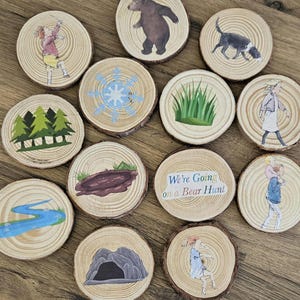 May include: A set of twelve circular wooden slices featuring illustrations from We're Going on a Bear Hunt, including a bear, a dog, a cave, a river, a snowstorm, grass, trees, and characters, with the title text printed on one wooden disc.