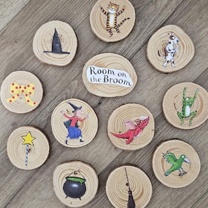 May include: A collection of eleven circular wooden slices featuring illustrations from Room on the Broom, including a witch, a cat, a dog, a frog, a bird, a dragon, a cauldron, a broomstick, a star wand, a hat, and a yellow polka dot bow.