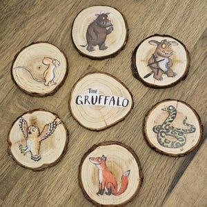May include: Seven circular wood slices featuring illustrations from The Gruffalo. One slice displays the text The Gruffalo, while the others show a mouse, a brown monster, a smaller creature, an owl, a snake, and a fox in various colours.