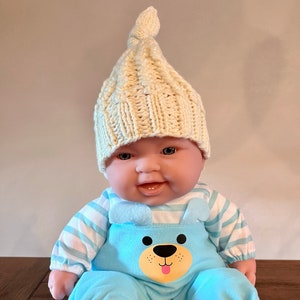 Puede incluir: Un muñeco de bebé de peluche con una camisa a rayas azul y blanca, un mono azul con una cara de oso en la parte delantera y un gorro de punto crema con un nudo en la parte superior.