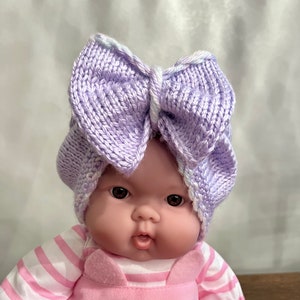 May include: A baby doll wearing a purple knitted bow headband and a pink and white striped shirt with a pink overall featuring a cat face.
