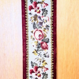 Könnte beinhalten: Eine antike Blumen-Wandteppich mit einem burgunderfarbenen Samtrücken und goldenen Verzierungen. Der Wandteppich zeigt ein Blumenmuster mit rosa, weißen und violetten Blumen. Der Wandteppich hängt an einer Holzwand mit einer dekorativen Metallglocke oben.