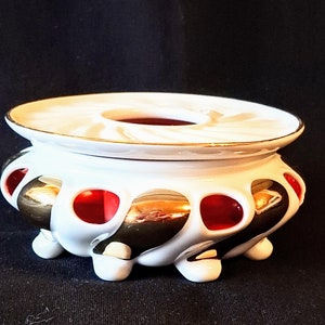 Könnte beinhalten: Weißer Keramik-Räucherofen mit gold-rotem Design. Der Räucherofen hat eine runde Oberseite mit einem Loch in der Mitte und einen Sockel mit drei Beinen.