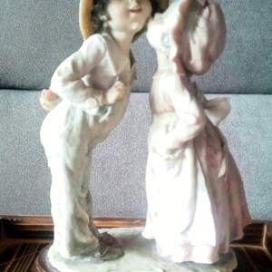 Könnte beinhalten: Eine Porzellanfigur von zwei Kindern, ein Junge mit Strohhut und ein Mädchen mit rosa Kleid, die sich küssen. Die Figur steht auf einem Holzsockel mit der Signatur des Künstlers.