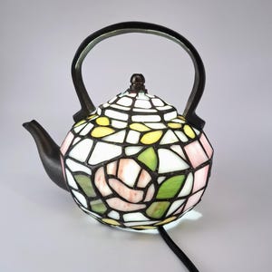 Könnte beinhalten: Eine Tiffany-Teekannenlampe mit schwarzem Griff, Ausguss und Sockel. Die Lampe hat ein Blumendesign mit rosa, grünen, gelben und weißen Glasteilen. Die Lampe ist mit einem schwarzen Kabel angeschlossen.