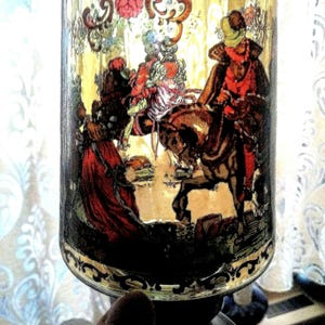 Könnte beinhalten: Eine klare Glasvase mit goldfarbenem Rand und einer bemalten Szene eines Mannes zu Pferd mit zwei Frauen. Die Szene ist in Rot-, Gelb- und Brauntönen gemalt.