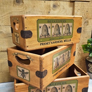 May include: Three wooden crates stacked, featuring the text "BEEKEEPERS HONEY & MEAD PRIORY GARDENS, WELLS". Each crate has a bee illustration and metal corner protectors. The crates are light brown with green and cream labels.