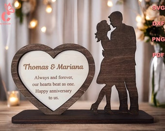 May include: A wooden heart-shaped frame with the text "Thomas & Mariana" and the message "Always and forever, our hearts beat as one. Happy anniversary to us."  A wooden silhouette of a couple embracing is standing next to the frame.