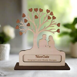 Puede incluir: Un recorte de madera de un &aacute;rbol con corazones en las ramas y una pareja de pie debajo. El &aacute;rbol est&aacute; sobre una base con el texto "NiceCuts Original Laser Ready Designs".