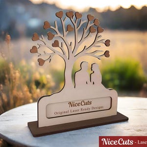 Puede incluir: Un recorte de madera de un &aacute;rbol con corazones como hojas y dos figuras de pie debajo. El &aacute;rbol est&aacute; sobre una base con el texto "Nice Cuts Original Laser Ready Designs".