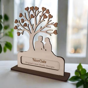Puede incluir: Corte de madera de una pareja de pie bajo un &aacute;rbol con corazones como hojas. El &aacute;rbol est&aacute; sobre una base con un letrero que dice "Nice Cuts Original Laser Ready Designs".
